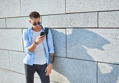 Genç bir adam, Avrupalı görünümlü, güneş gözlüklü ve siyah sırt çantalı. Duvarın yanında duruyor ve kulaklıkla müzik dinlerken telefona bakıyor..