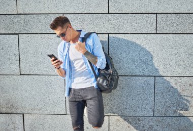 Genç bir adam, Avrupalı görünümlü, güneş gözlüklü ve siyah sırt çantalı. Duvarın yanında duruyor ve kulaklıkla müzik dinlerken telefona bakıyor..