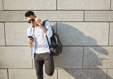 Genç bir adam, Avrupa görünümlü, güneş gözlüklü ve siyah sırt çantalı. Duvarın yanında duruyor, telefonu elinde tutuyor ve çerçeveye bakıyor..