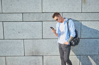Genç bir adam, Avrupalı görünümlü, güneş gözlüklü ve siyah sırt çantalı. Duvarın yanında duruyor ve kulaklıkla müzik dinlerken telefona bakıyor..