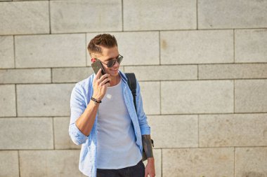 Genç bir adam, Avrupa görünümlü, güneş gözlüklü ve siyah sırt çantalı. Şehir caddesinde telefonla konuşuyor..