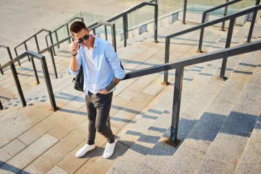 Siyah sırt çantalı, güneş gözlüklü genç bir adam merdivenlerde durup korkuluklara yaslanıyor..
