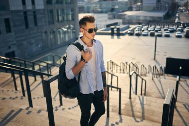 Güneş gözlüklü genç adam merdivenlerden siyah bir sırt çantasıyla çıktı.