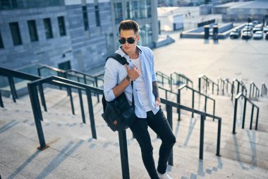 Siyah sırt çantalı, güneş gözlüklü genç bir adam merdivenlerde durup korkuluklara yaslanıyor..