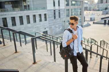 Siyah sırt çantalı, güneş gözlüklü genç bir adam merdivenlerde durup korkuluklara yaslanıyor..