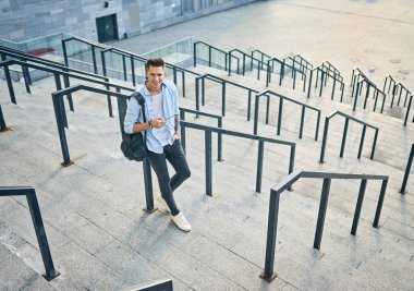 Güneş gözlüklü ve siyah sırt çantalı genç bir adam. Basamaklarda durup gülümsüyor.