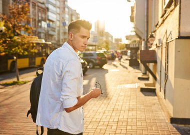Elinde güneş gözlüğü olan siyah bir sırt çantalı genç bir adam..