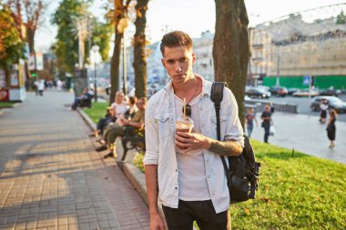 Genç Avrupalı bir adam şehir caddesinde kahve içiyor. Sırt çantalı, güzel bir gün batımı ışığı var..