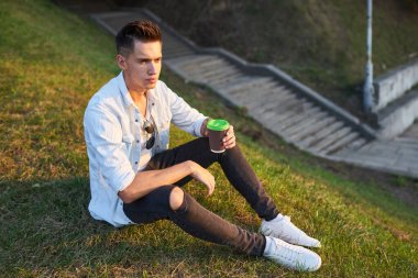 Genç Avrupalı bir adam şehir caddesinde kahve içiyor. Sırt çantalı, güzel bir gün batımı ışığı var..