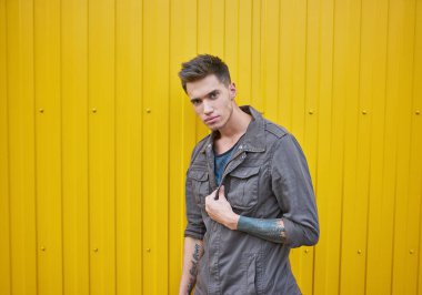 Kolunda kot ceketli bir dövme olan genç moda adamı, sarı duvarın yanında duruyor.