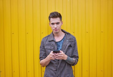 Genç bir adam sarı duvarın yanında duruyor, telefonu elinde tutuyor ve çerçeveye bakıyor..