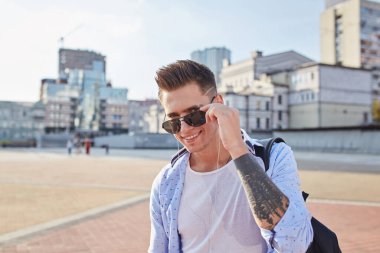 Güneş gözlüklü, siyah sırt çantalı, şehir caddesinde yürüyen şık bir genç.