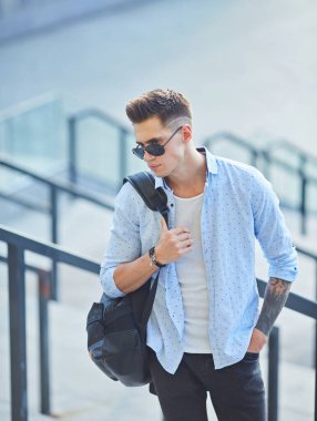 Siyah sırt çantalı, güneş gözlüklü genç bir adam merdivenlerde durup korkuluklara yaslanıyor..