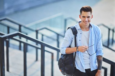 Güneş gözlüklü ve siyah sırt çantalı genç bir adam. Basamaklarda durup gülümsüyor.