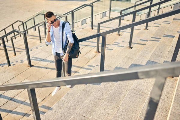 A young man with a black backpack standing on a city street holding sunglasses in his hands.