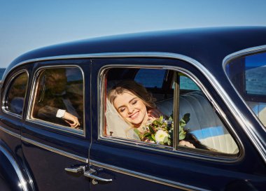 Lüks gelinlikli güzel gelin eski arabanın içinde oturuyor ve elinde düğün buketi tutuyor.