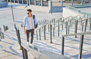 Güneş gözlüklü genç adam merdivenlerden siyah bir sırt çantasıyla çıktı.