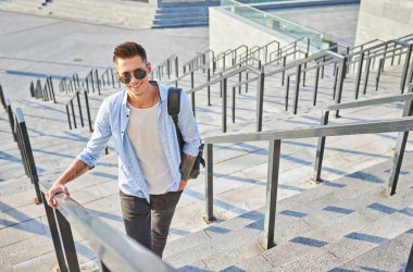 Güneş gözlüklü ve siyah sırt çantalı genç bir adam. Basamaklarda durup gülümsüyor.