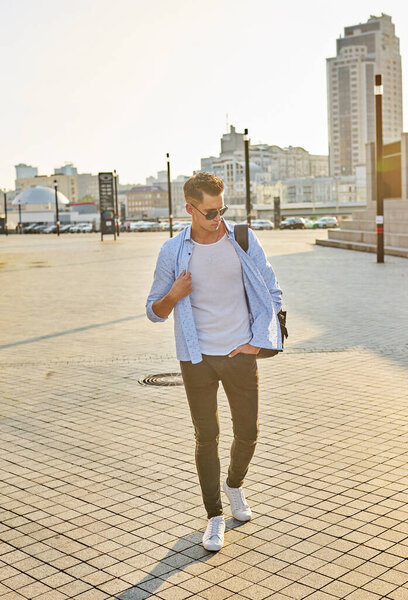 Fashionable young man in sunglasses with a black backpack walking along a city street