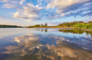 Yazın gün batımında güzel nehir kıyısında. Göl, yeşil ağaçlar ve çimenler ile renkli manzara, mavi gökyüzü çok renkli bulutlar ve suya yansıyan turuncu güneş ışığı. Doğa. Canlı bir manzara.