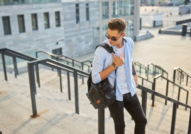 Siyah sırt çantalı, güneş gözlüklü genç bir adam merdivenlerde durup korkuluklara yaslanıyor..