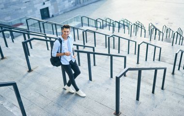 Siyah sırt çantalı, güneş gözlüklü genç bir adam merdivenlerde durup korkuluklara yaslanıyor..