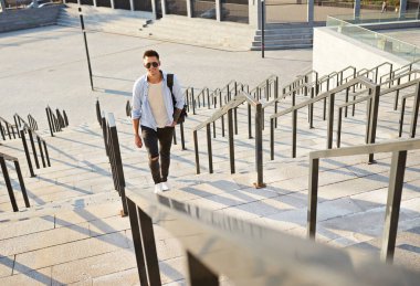 Güneş gözlüklü ve siyah sırt çantalı genç bir adam. Basamaklarda durup gülümsüyor.