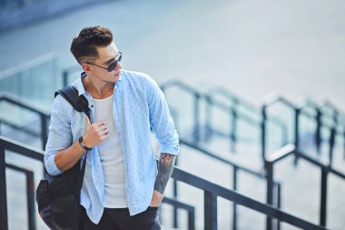 Siyah sırt çantalı, güneş gözlüklü genç bir adam merdivenlerde durup korkuluklara yaslanıyor..