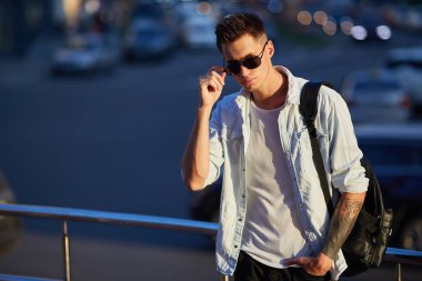 Elinde güneş gözlüğü olan siyah bir sırt çantalı genç bir adam..