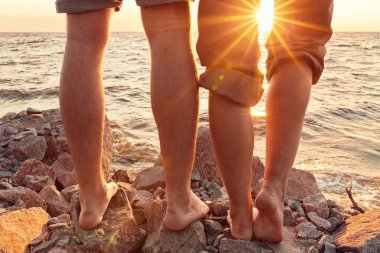 Bacakları sahilde. Ayak spası. Gün batımında sahilde sarılıp öpüşen genç bir çift. İki sevgili, erkek ve kadın, suyun yanında yalınayak. Yaz aşkı.