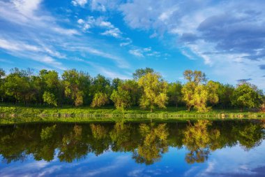 Yazın gün batımında güzel nehir kıyısında. Göl, yeşil ağaçlar ve çimenler ile renkli manzara, mavi gökyüzü çok renkli bulutlar ve suya yansıyan turuncu güneş ışığı. Doğa. Canlı bir manzara.
