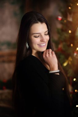 Genç esmer, neşeli bir ruh hali içinde, Noel ağacının yanında siyah bir elbise içinde.