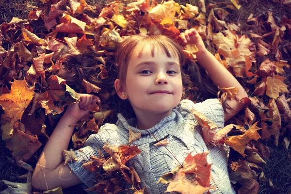 Adorable little girl in a sweater lying on autumn leaves in the park — Stock Photo, Image
