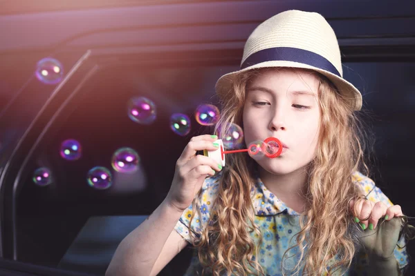 Uma menina adorável em um chapéu deixa bolhas de sabão no carro — Fotografia de Stock