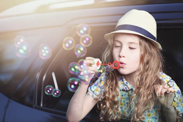 Petite fille mignonne dans un chapeau laisse bulles de savon dans la fenêtre de la voiture — Photo