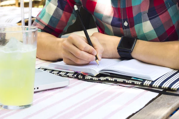 Detailní záběr rukou psát něco v deníku poznámkového bloku. Stylový muž pracuje v kavárně s limonade. — Stock fotografie