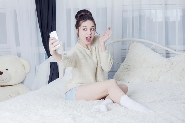 Surprised young woman taking selfie on the phone on the bed — Stock Photo, Image