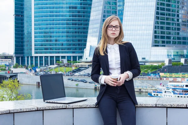 Attraktiva affärskvinna med kaffe och en laptop på bakgrund av skyskrapor — Stockfoto