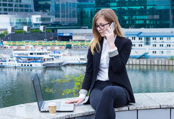 En forretningskvinne som skriver på en bærbar datamaskin og snakker i telefonen i bakgrunnen av skyskrapere – stockfoto