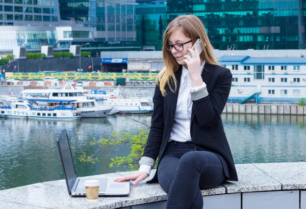 Mujer de negocios escribiendo en el ordenador portátil y hablando por teléfono en el fondo de los rascacielos — Foto de Stock