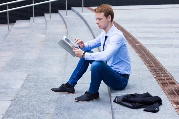 Joven empresario sentado en la calle cerca del centro de negocios y las obras en la tableta — Foto de Stock