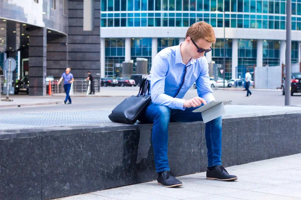 Young businessman sitting in the street near the business center and the works on the tablet — Stock Photo, Image