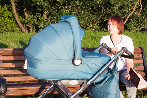 Moeder lezen van een boek op een bankje met haar baby in een wandelwagen in het park Rechtenvrije Stockfoto's