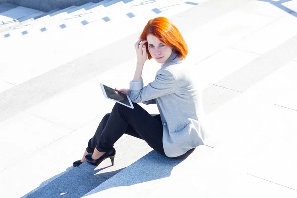 Mulher ruiva sentada nas escadas e lendo algo com um tablet no dia ensolarado — Fotografia de Stock