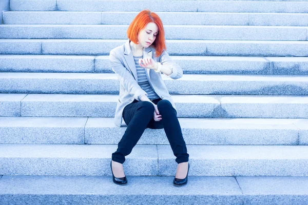 Руда приваблива жінка сидить на вулиці на сходах і дивиться час — стокове фото