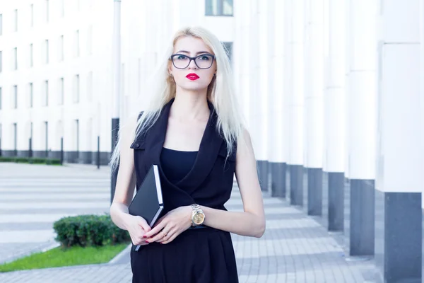 Интеллигентная блондинка с красными губами и блокнотом ждет кого-то на фоне бизнес-центра — стоковое фото