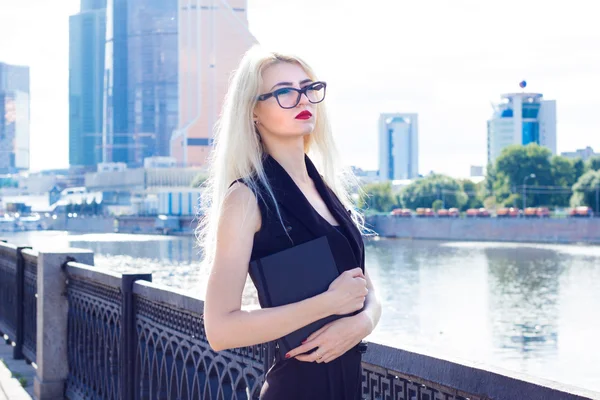 Умная блондинка с красными губами и блокнотом смотрит вдаль на фоне небоскребов — стоковое фото