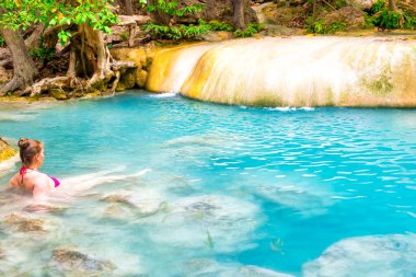 Pembe bikinili kızıl saçlı genç kadın tropik gölün zümrüt mavisi sularında yüzüyor. Erawan Ulusal Parkı, Kanchanaburi, Tayland