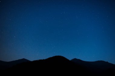 Karanlık mavi gökyüzünün altında parlak yıldızlarla dolu bir dağ silueti.
