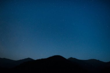 Karanlık mavi gökyüzünün altında parlak yıldızlarla dolu bir dağ silueti.